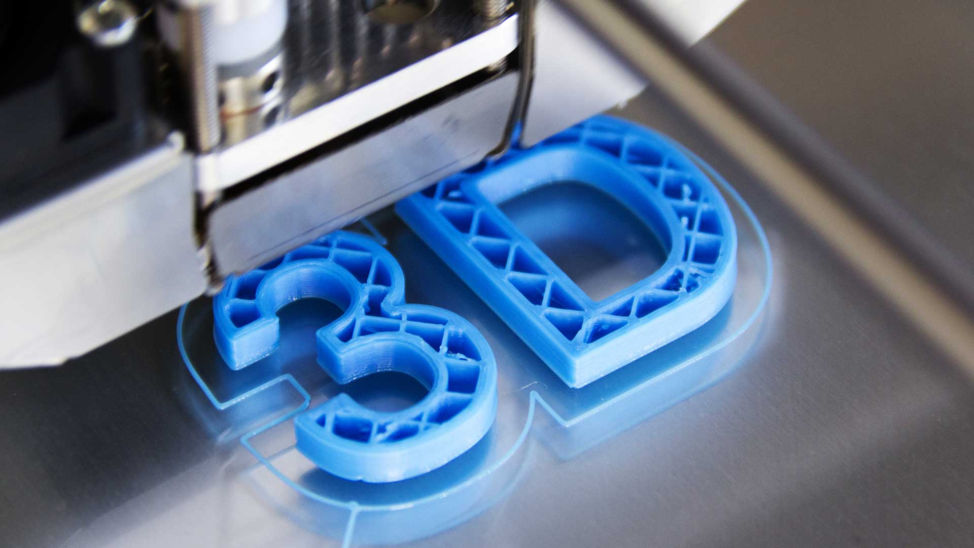 Drukarka 3D w trakcie drukowania niebieskich liter &bdquo;3D&rdquo; z filamentu, ilustrująca usługę druku 3D