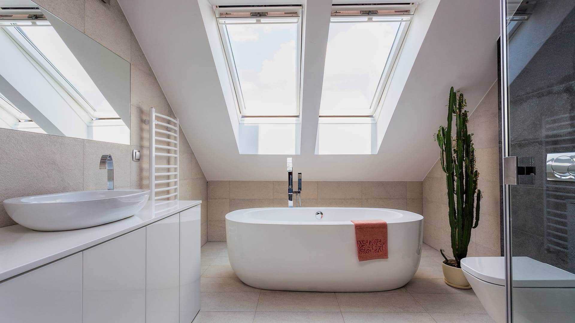 Jasna łazienka na poddaszu ze skosami oraz oknami dachowymi i białą wanną wolnostojącą
