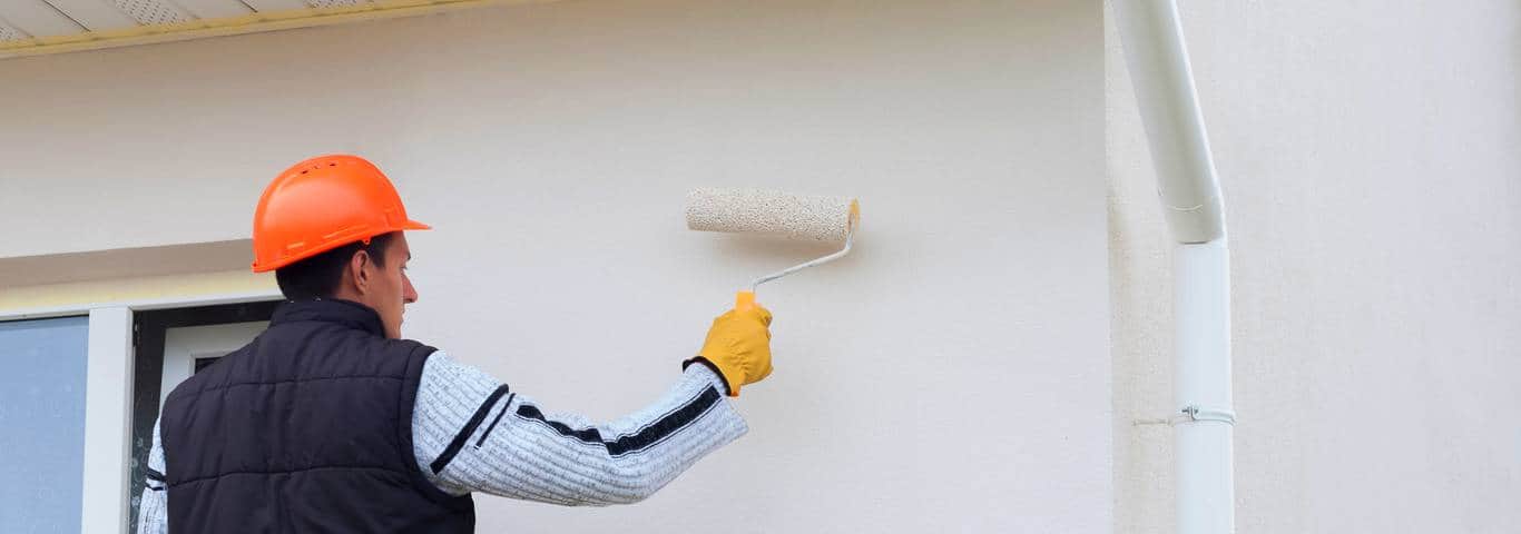 malarz w kasku ochronnym maluje elewację domu wałkiem