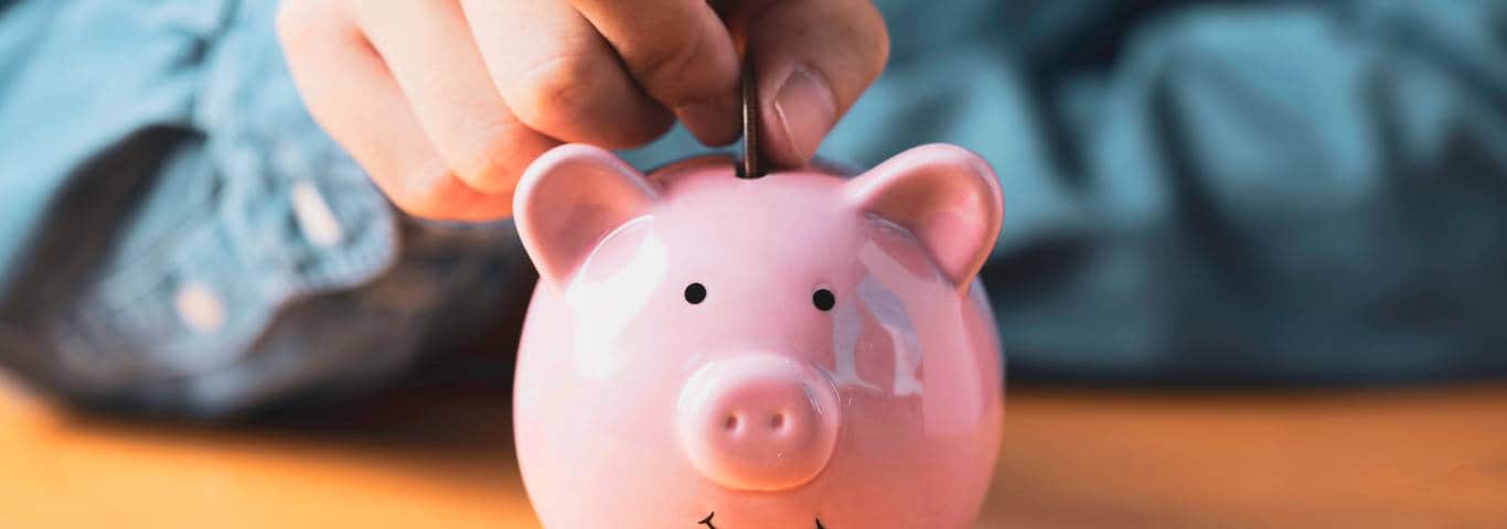 Ręka wkładająca monetę do różowej skarbonki – symbol inwestycji w komfort i oszczędność