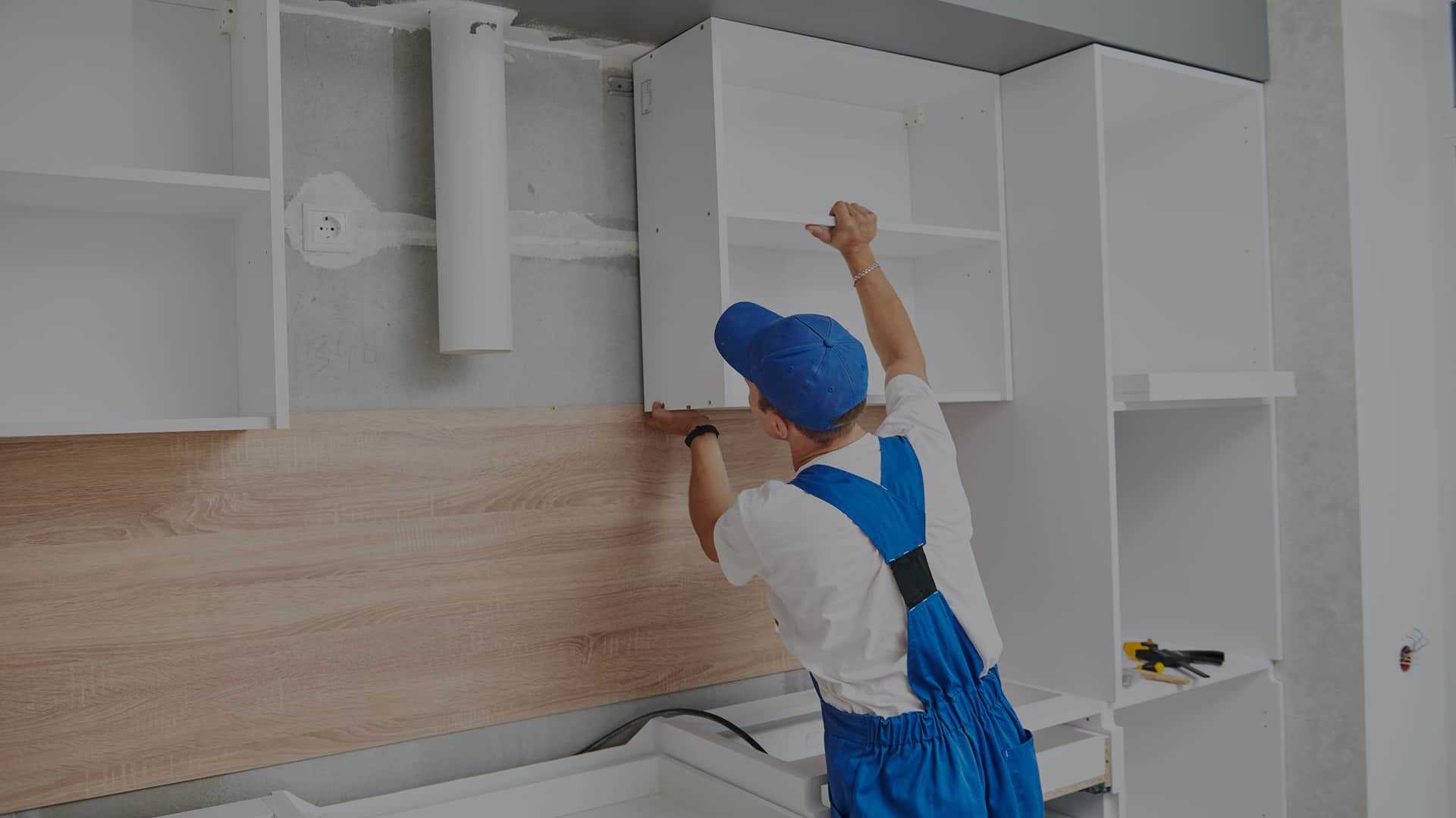 Stolarz w niebieskim kombinezonie montuje białą szafkę kuchenną na ścianie