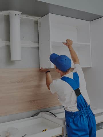 Stolarz w niebieskim kombinezonie montuje białą szafkę kuchenną na ścianie