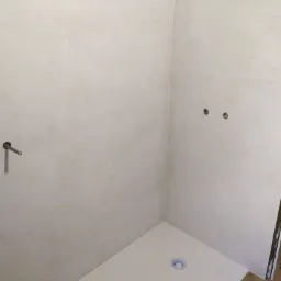 Płytki ścienne imitujące kamień w jasnym odcieniu szarości, zamontowany brodzik z odpływem oraz wystające przyłącza wodne i kanalizacyjne w narożniku łazienki w trakcie prac wykończeniowych.