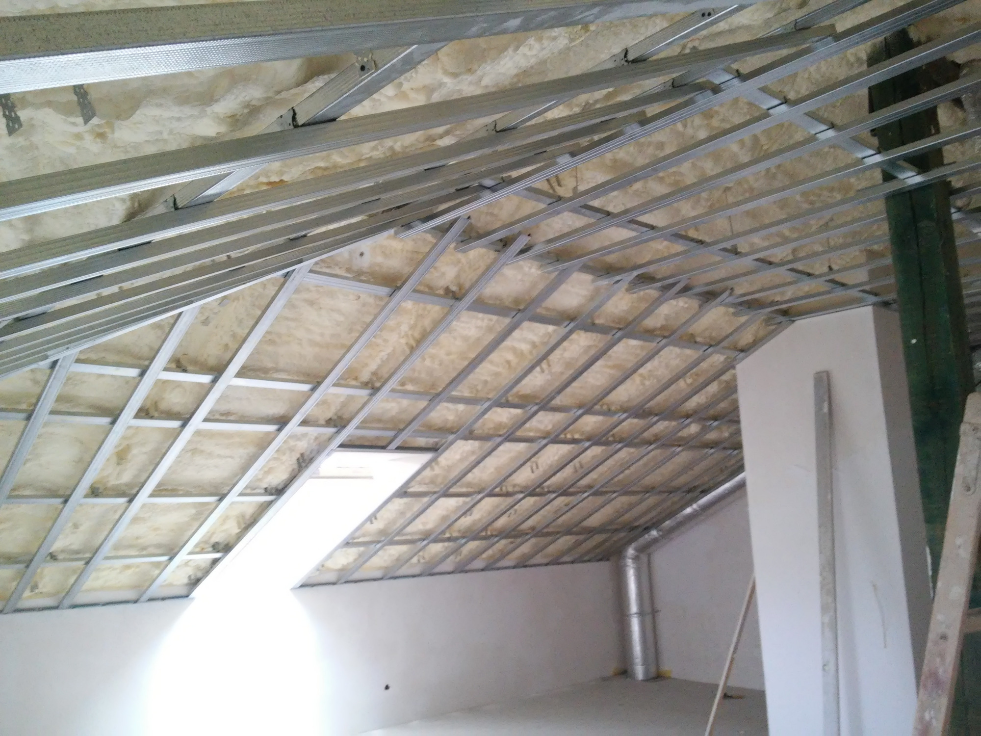 Szkielet sufitu podwieszanego z izolacją z wełny mineralnej w poddaszu, widoczne profile metalowe i fragment instalacji wentylacyjnej.