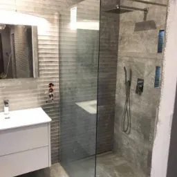 Nowoczesna łazienka z szarą, teksturowaną glazurą, kabiną prysznicową typu walk-in z deszczownicą, białą szafką pod umywalką i minimalistyczną armaturą.