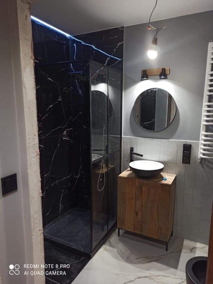 Nowoczesna łazienka z czarną kabiną prysznicową z marmurowym wzorem, drewnianą szafką pod umywalką i okrągłym lustrem, widoczne niedokończone wykończenie ściany po lewej stronie.