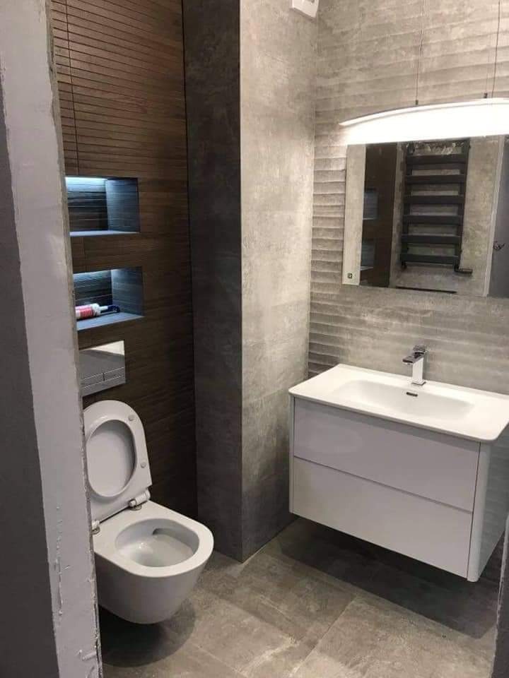 Nowoczesna łazienka w szaro-brązowej tonacji: wisząca toaleta z otwartą klapą, biała szafka z umywalką, lustro z oświetleniem i drewniane panele z wnękami podświetlanymi LED.