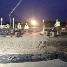 Proces wylewania posadzki na placu budowy nocą, widoczni pracownicy w odblaskowych kamizelkach i kaskach, maszyny do rozprowadzania betonu, zbrojenie w siatce.