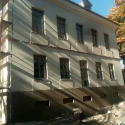 Elewacja budynku w trakcie tynkowania, widoczne rusztowania, część elewacji zabezpieczona siatką, jesienne drzewa w tle.