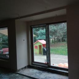 Rotis - Surowe ściany w trakcie prac, widoczne kable wystające z sufitu, okna z widokiem na ogród z domkiem dla dzieci.