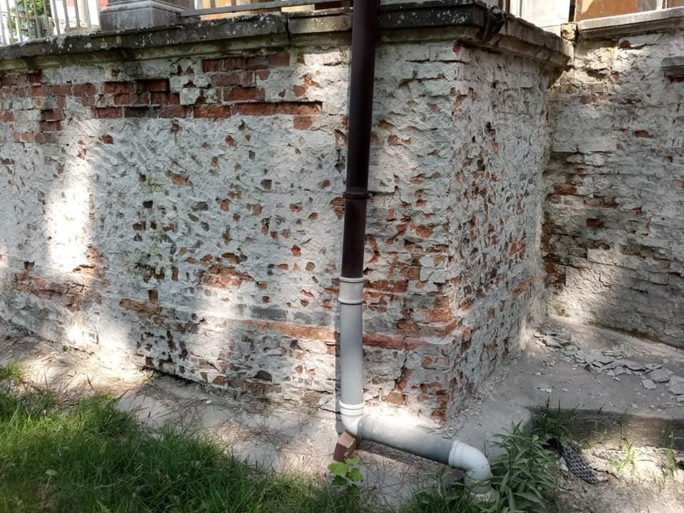 Fragment zniszczonej ceglanej elewacji z resztkami starego tynku, widoczny róg budynku i rura spustowa, teren wokół zarośnięty trawą, Wrocław.