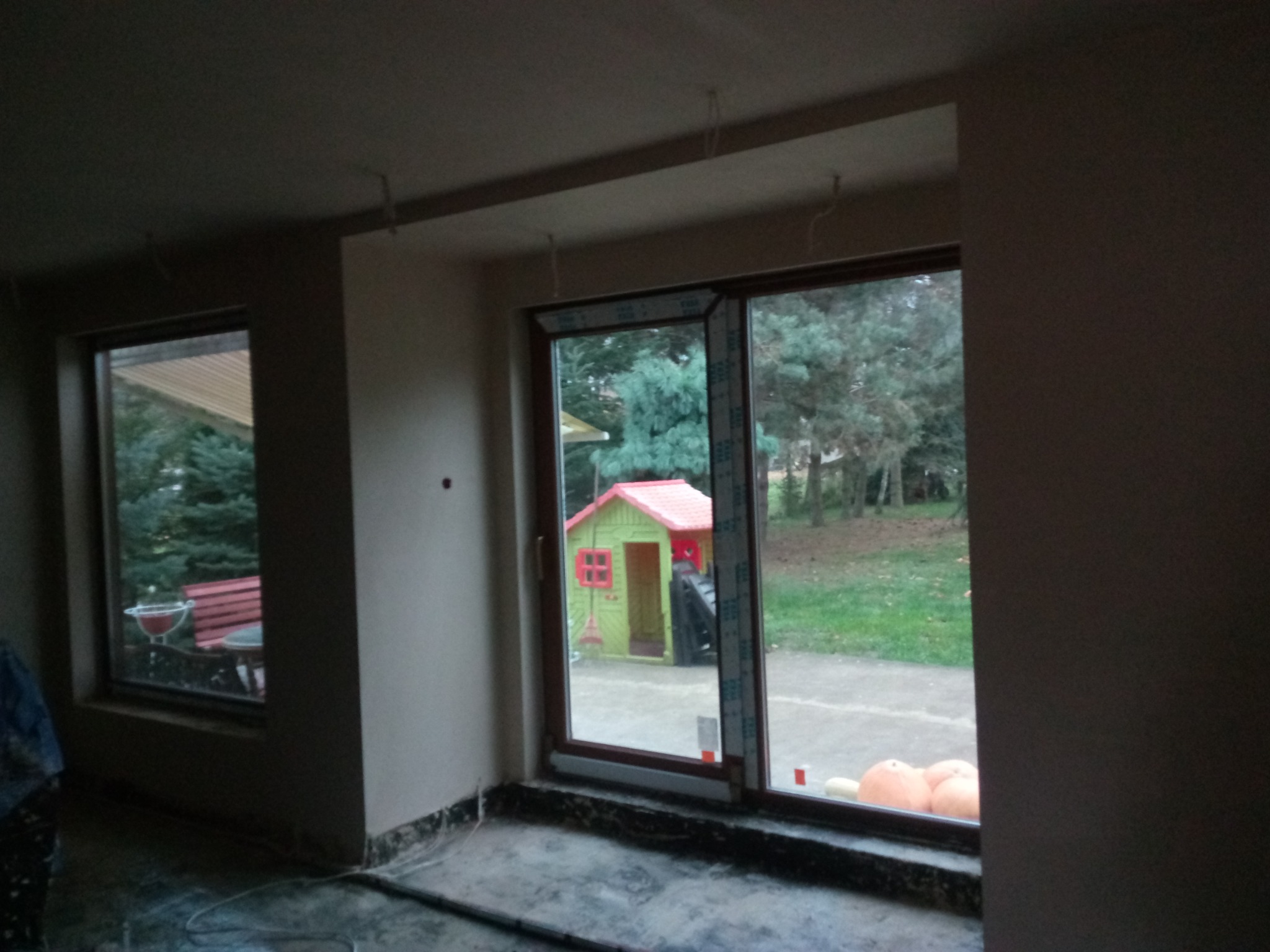 Surowe ściany w trakcie prac, widoczne kable wystające z sufitu, okna z widokiem na ogród z domkiem dla dzieci.