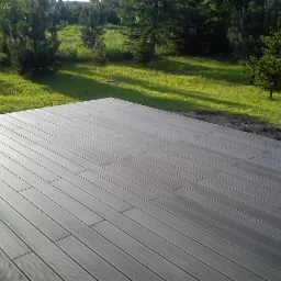 Nowoczesny taras z desek kompozytowych w odcieniach szarości, widok z niskiej perspektywy na tle zielonego trawnika i drzew iglastych w słoneczny dzień.