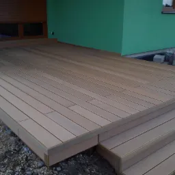 Gotowy taras z desek kompozytowych w odcieniach brązu, z niskimi schodami, na tle zielonej ściany budynku z brązowymi roletami okiennymi, widoczne kamienie pod konstrukcją.