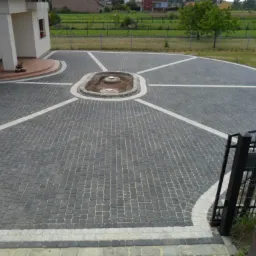 Obszerny podjazd z ciemnoszarej kostki, z centralnym, owalnym klombem i białymi liniami rozchodzącymi się promieniście, prowadzący do domu z kolumnami i schodami.