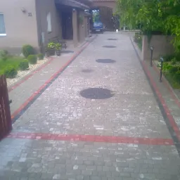 Ułożona z kostki brukowej podjazd do domu z numerem 21, widoczne studzienki kanalizacyjne, oświetlenie ogrodowe i fragment drewnianego ogrodzenia.