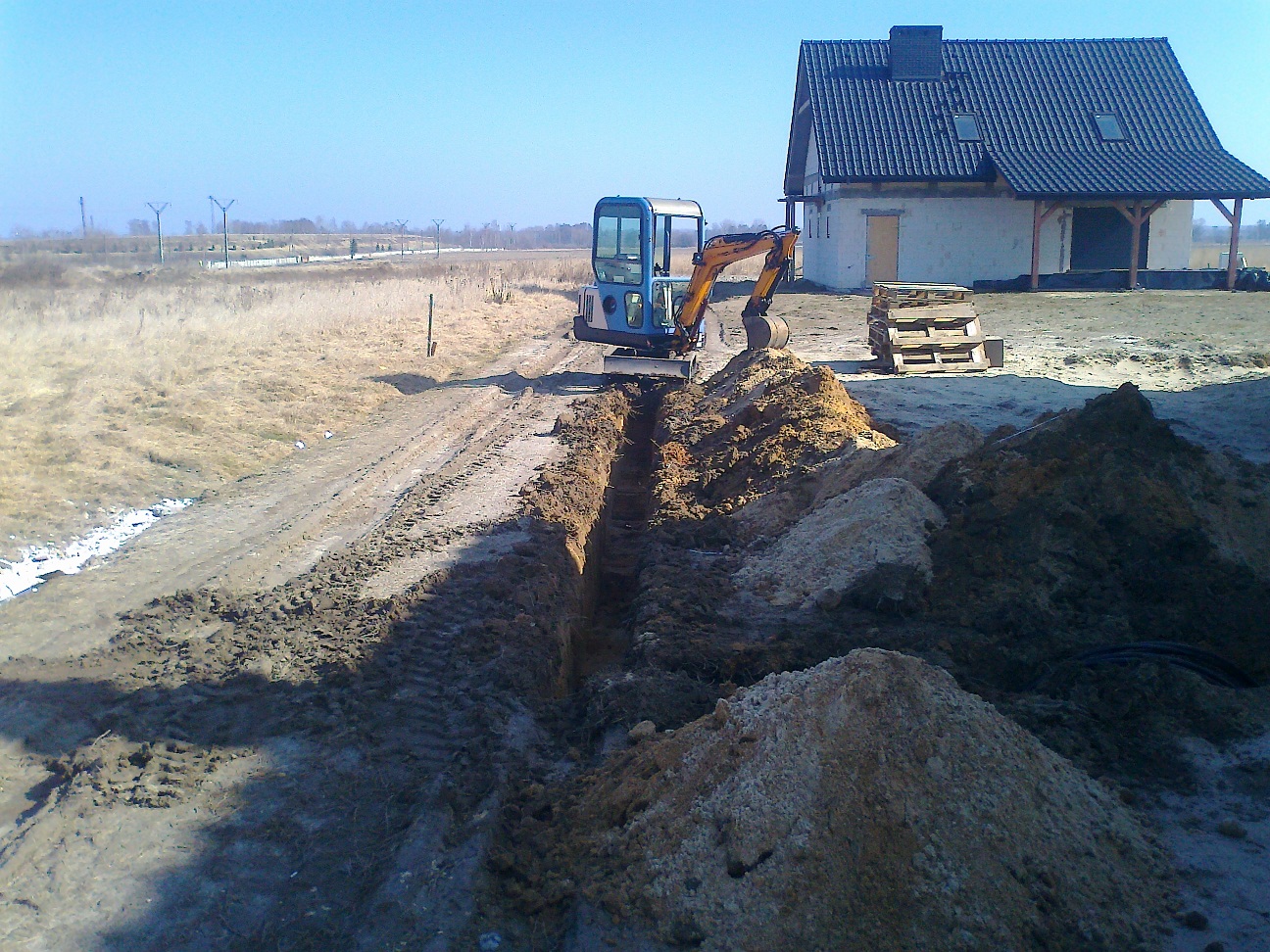 Niebieska minikoparka tworzy wąski wykop w ziemi na tle budowanego domu z ciemnym dachem i stertą palet.