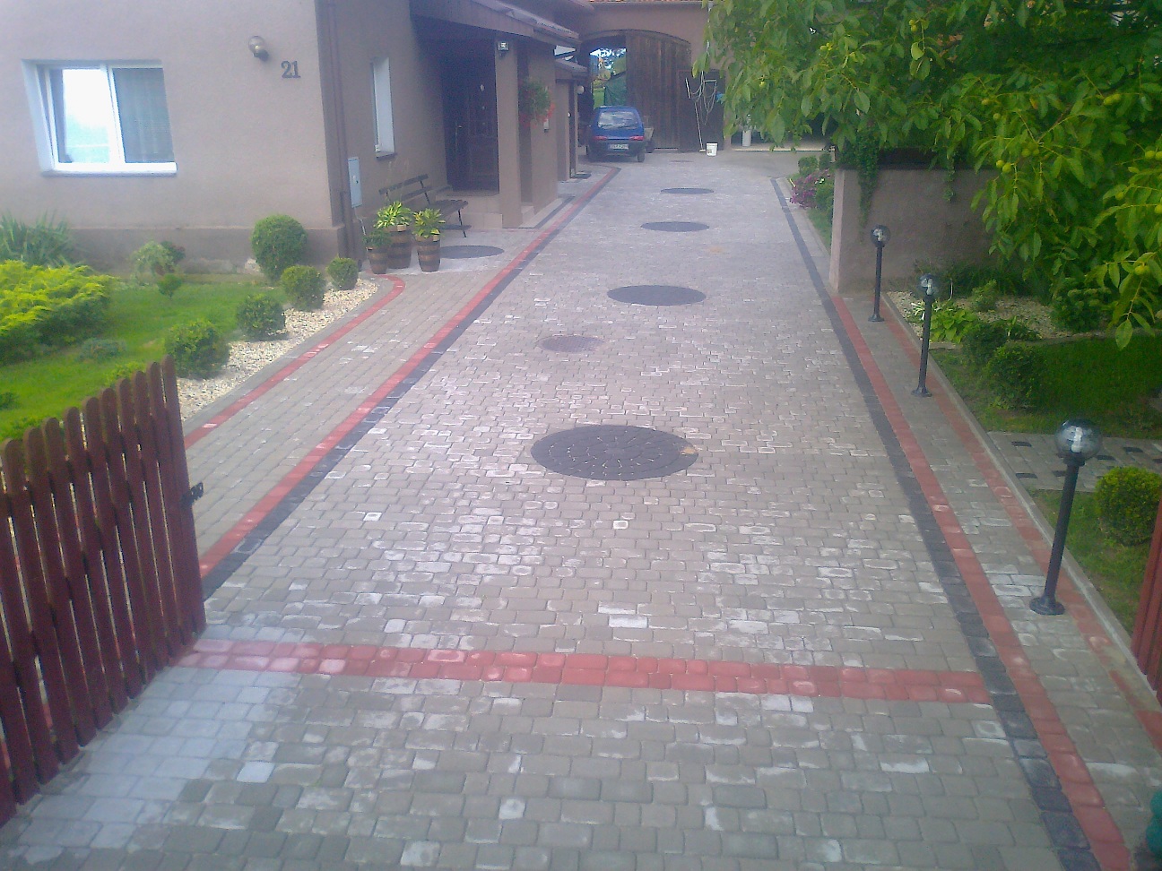 Ułożona z kostki brukowej podjazd do domu z numerem 21, widoczne studzienki kanalizacyjne, oświetlenie ogrodowe i fragment drewnianego ogrodzenia.