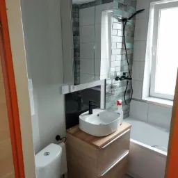 Umeblowana łazienka z białą ceramiką, umywalką nablatową na drewnianej szafce, wanną i armaturą w kolorze czarnym, wykończona płytkami w odcieniach bieli i turkusu.