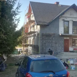 Dom w trakcie ocieplania styropianem, widoczne rusztowania, zaparkowany niebieski samochód Toyota Yaris na trawniku.