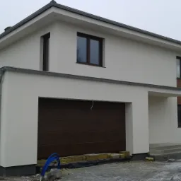 Nowoczesny, dwukondygnacyjny dom z płaskim dachem, brązowymi oknami i bramą garażową w kolorze drewna. Na pierwszym planie widoczne materiały budowlane i niebieskie rury.