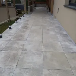 Wykończony taras z płyt wentylowanych w odcieniach szarości, z widocznymi wspornikami regulowanymi, drewnianą pergolą i elewacją budynku w tle.