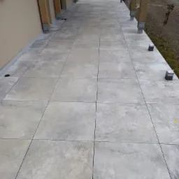 Wykończony taras z szarych, kwadratowych płyt betonowych, wsparty na drewnianych słupach z metalowymi łącznikami, z widocznymi elementami wykończeniowymi na obrzeżach.