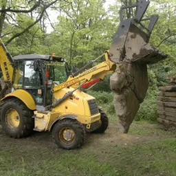 Żółta koparko-ładowarka New Holland podnosi duży, nieregularny blok ziemi i kamieni w zalesionym terenie, obok sterty płyt.
