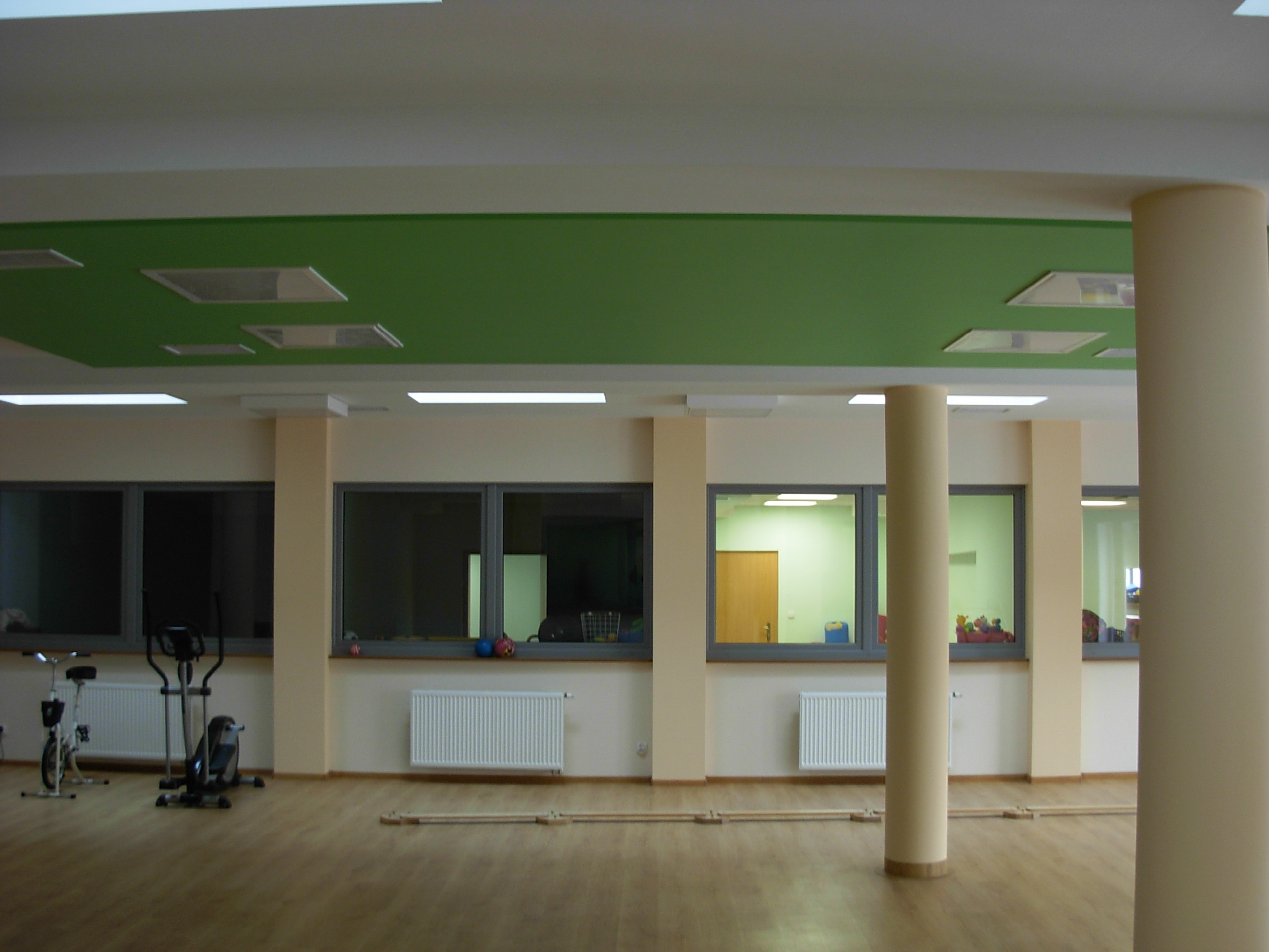 Jasne wnętrze sali z drewnianą podłogą, grzejnikami pod oknami i kolumnami, na suficie zielony panel z wbudowanym oświetleniem.
