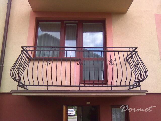 Kuta balustrada balkonowa z dekoracyjnymi elementami w kształcie ślimaków, zamontowana na balkonie z brązową ramą okienną i beżowo-różową elewacją budynku.