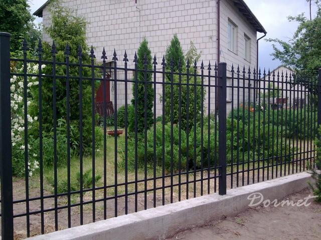 Czarne, metalowe przęsła ogrodzeniowe z ozdobnymi grotami na tle zadbanego ogrodu i domu z białej cegły.
