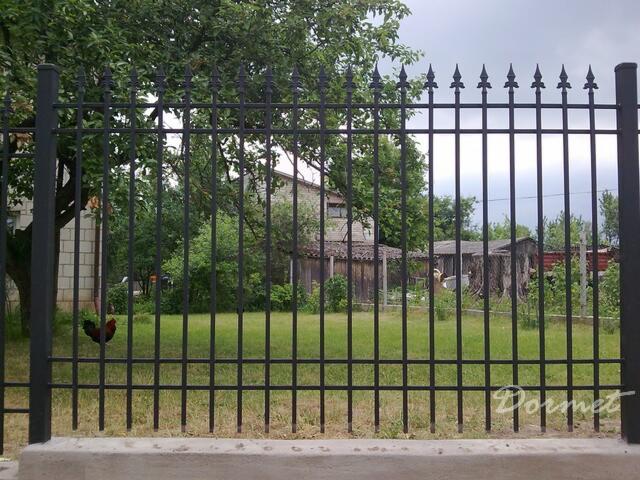 Czarne, metalowe przęsło ogrodzeniowe z grotami na szczycie, widok na zielony trawnik, kurnik i budynek w tle, na trawniku widać czarnego koguta.