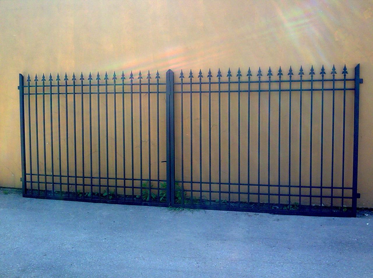 Dwuskrzydłowa brama z kutego żelaza, zakończona grotami, zamontowana przy żółtej ścianie, z widoczną furtką i niewielką roślinnością u podstawy.