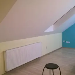 Pokój na poddaszu po remoncie: skośny sufit, niebieska ściana, drewniana podłoga, biały grzejnik, krzesło. Widoczne gniazdka elektryczne.
