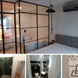 Sypialnia z widokiem na łazienkę oddzieloną szklaną ścianą z czarnymi profilami, widoczna wanna wolnostojąca, umywalka w kolorze koralowym, na podłodze marmur.