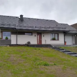 Dom jednorodzinny z szarą dachówką w Trzebini, widoczny z perspektywy trawnika. Elewacja w odcieniach bieli i szarości, z elementami dekoracyjnymi z kamienia przy oknach. Nowoczesne schody prowadzą...