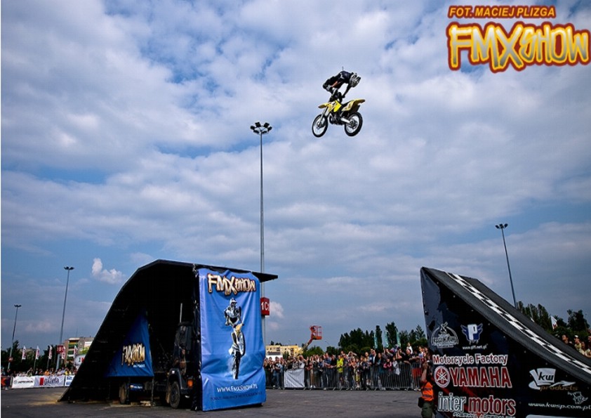 Żółty motocykl z motocyklistą wykonuje skok w powietrzu podczas pokazu FMX Show, w tle widoczna rampa, tłum widzów za barierkami oraz błękitne niebo z chmurami.