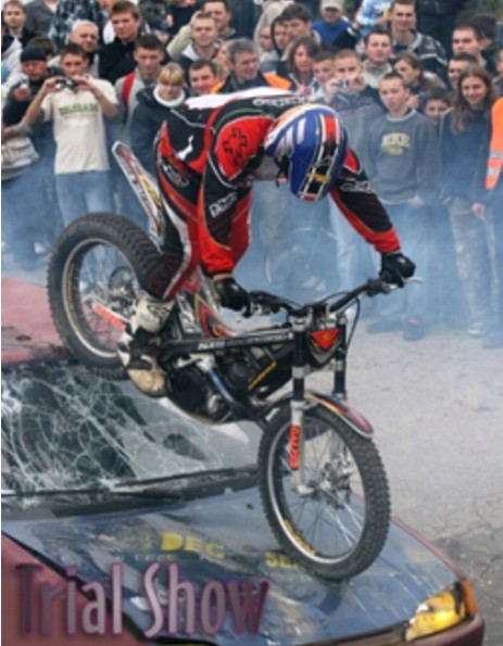 Motocyklista w kasku i kombinezonie wykonuje akrobacje na motocyklu trialowym, stojąc na masce zniszczonego samochodu na tle publiczności.