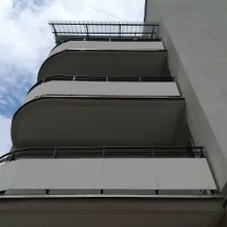 Trzy zaokrąglone balkony z metalowymi balustradami i białymi panelami, widok z dołu na tle pochmurnego nieba.