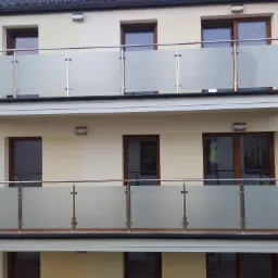 Dwa balkony z metalowymi balustradami i mlecznym szkłem, widoczne z zewnątrz budynku o jasnej elewacji i ciemnych oknach.