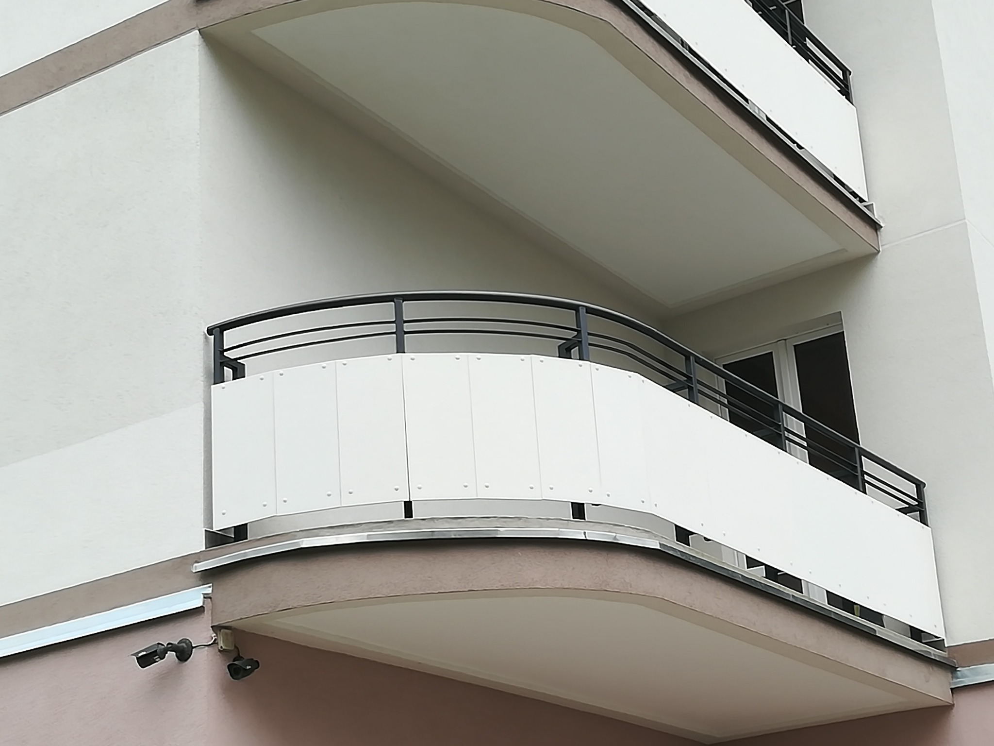 Nowoczesny balkon z metalową balustradą i białymi panelami, widok z dołu, z dwoma kamerami monitoringu zamontowanymi pod spodem balkonu.