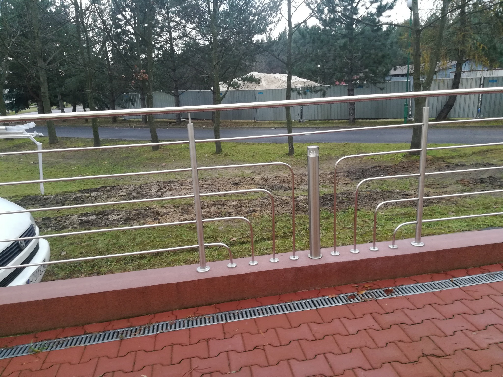 Metalowa balustrada ochronna z poprzecznymi prętami i pionowymi słupkami, zamontowana na niskim murku z czerwonej cegły, widok z perspektywy chodnika wyłożonego czerwoną kostką.