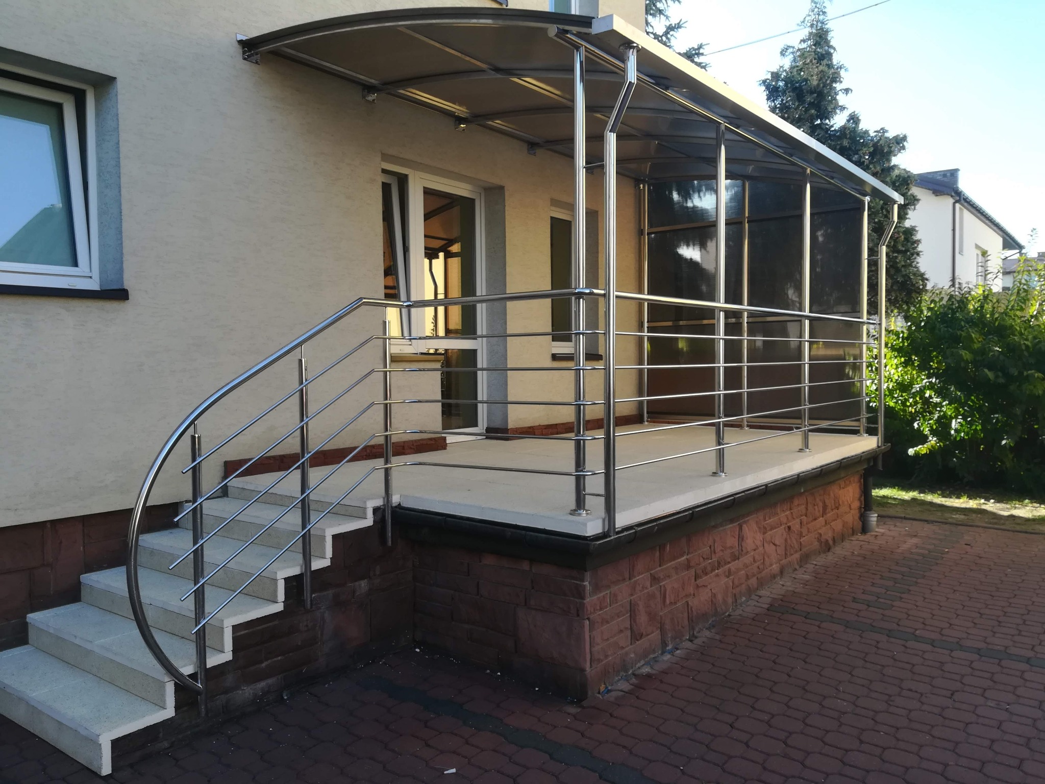 Zewnętrzne schody betonowe z metalową balustradą i zadaszeniem z poliwęglanu, prowadzące do wejścia budynku z jasną elewacją i oknem.