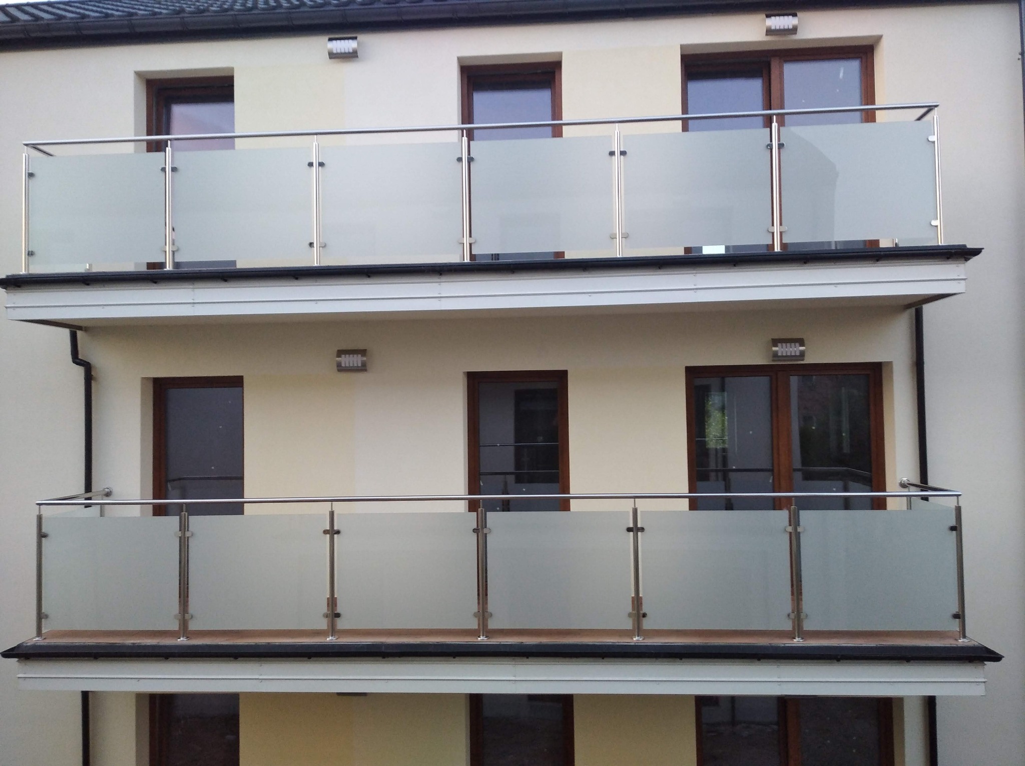 Dwa balkony z metalowymi balustradami i mlecznym szkłem, widoczne z zewnątrz budynku o jasnej elewacji i ciemnych oknach.