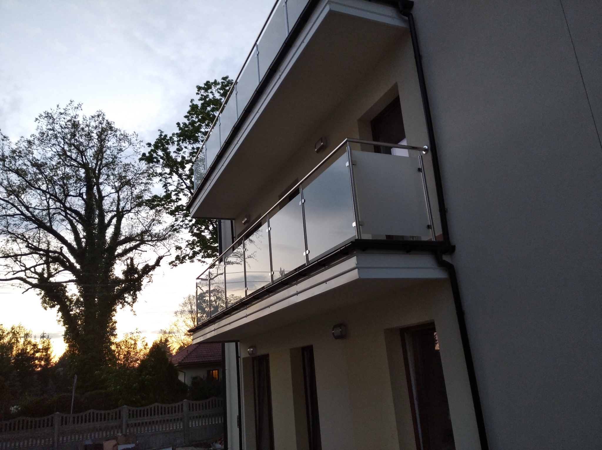 Dwa balkony z metalowo-szklanymi balustradami na tle zachodzącego słońca i dużego drzewa.