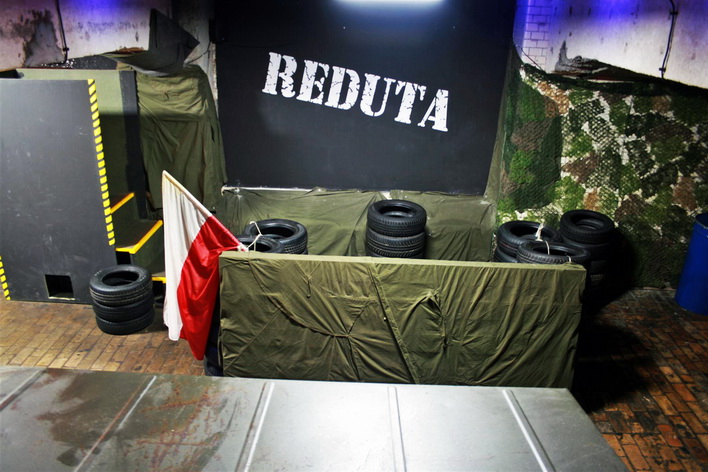 Przygotowana arena do rozgrywek taktycznych z przeszkodami, oponami i flagą Polski, podświetlona niebieskim światłem, z napisem 'Reduta' na czarnym tle.