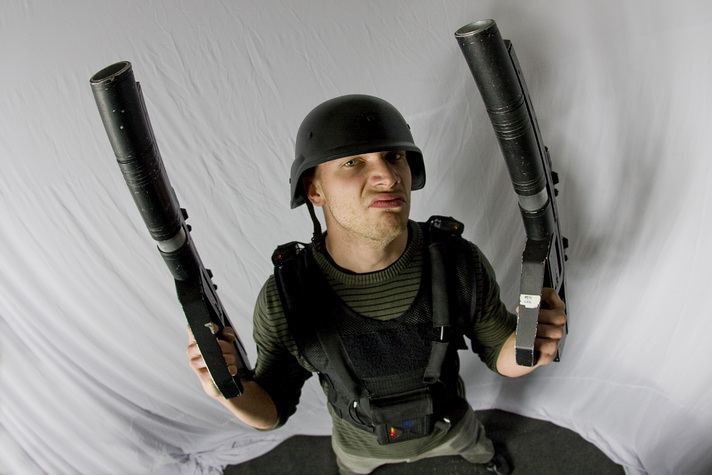 Mężczyzna w hełmie i kamizelce taktycznej, z grymasem na twarzy, trzymający w obu rękach duże, czarne karabiny na tle białego materiału.