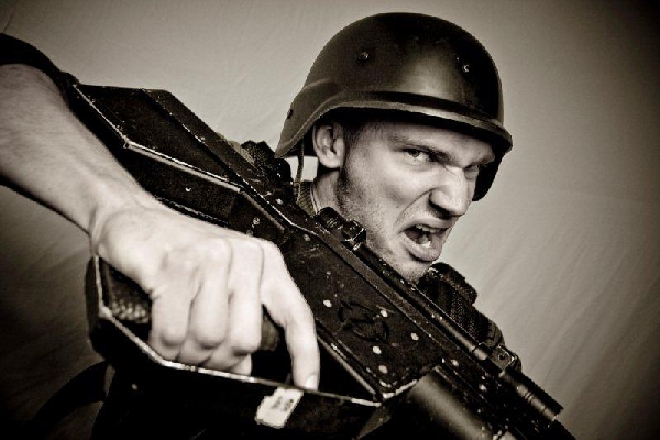 Mężczyzna w hełmie, z grymasem na twarzy, trzymający w rękach karabin, skierowany lufą w stronę widza, w sepiowej tonacji.