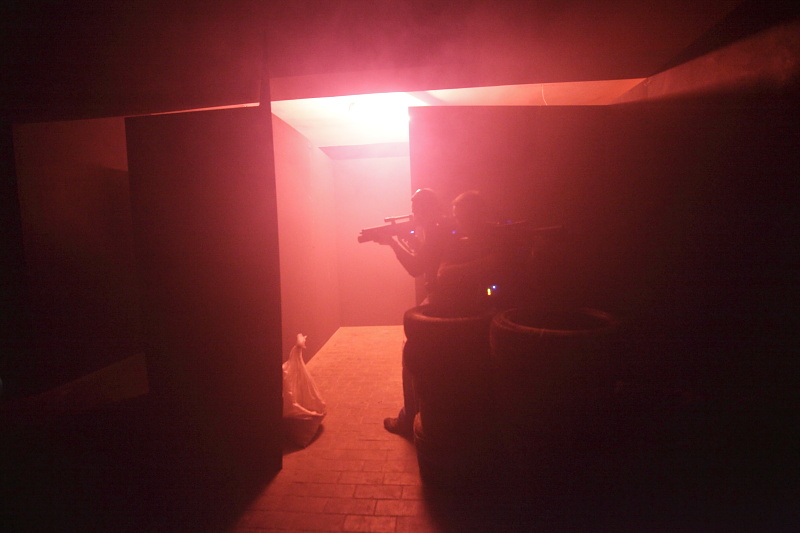 Dwie osoby w ciemnym labiryncie oświetlonym czerwonym światłem, trzymające karabiny do laserowego paintballa, kryjące się za stertą opon.