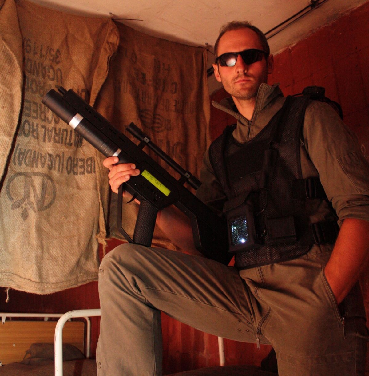 Mężczyzna w kamizelce taktycznej i okularach przeciwsłonecznych, siedzący na łóżku z futurystycznym karabinem w ręku, pozujący na tle worków jutowych w scenerii przypominającej wnętrze bazy.